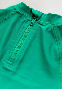 Vêtement zippé vert avec un col haut, doté d'un tissu lisse et extensible et de coutures à plat le long des coutures. Le zip est central et visible.