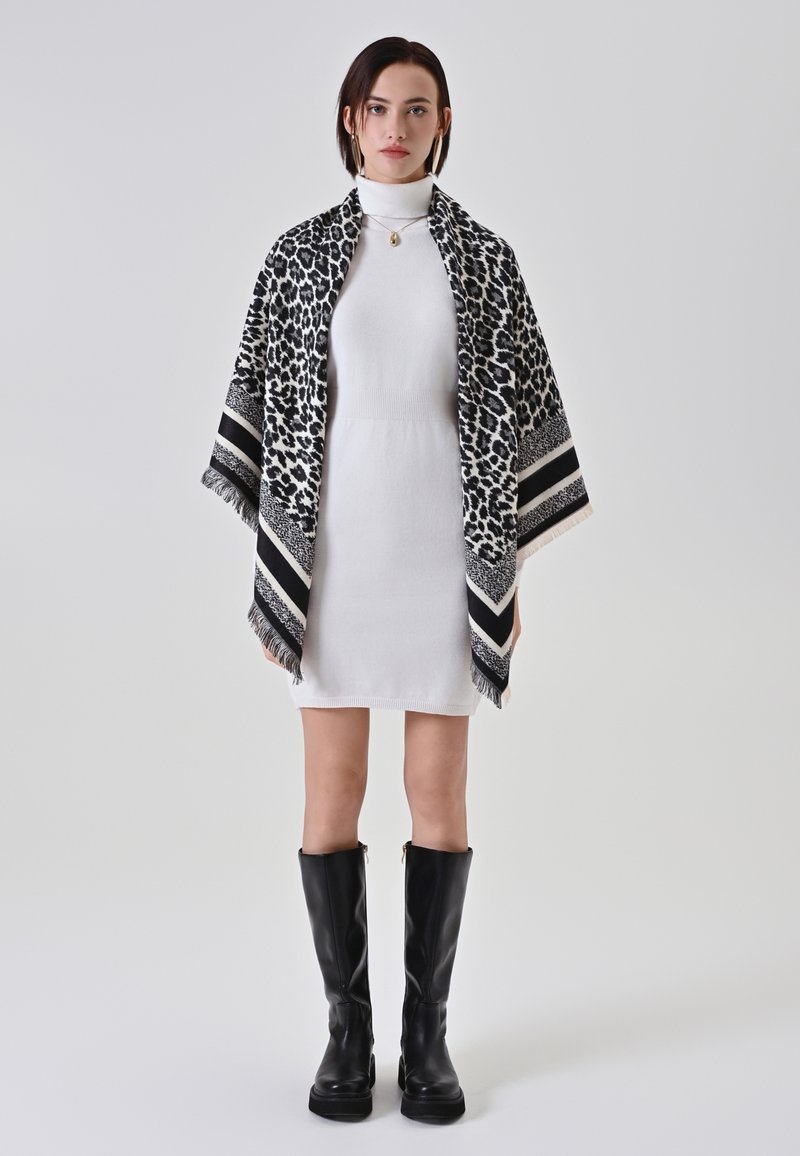 Châle imprimé léopard noir et blanc avec des bords à franges et des accents rayés, porté sur une robe blanche avec des bottes noires à longueur de cheville.