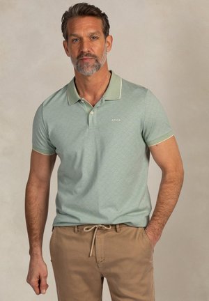 Man met een grijsspoorrige baard draagt een lichtgroen polo met patroon en beige broek, staand tegen een neutrale achtergrond.