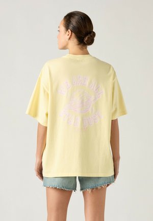 Junge Frau mit Dutt, die ein blassgelbes oversized T-Shirt mit der Aufschrift „We Are All Star Dust“ und Planeten-Grafik auf dem Rücken trägt, kombiniert mit Jeansshorts.