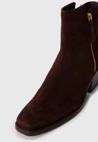 Bottine en daim marron avec un bout carré et une fermeture éclair latérale. Dotée d'un talon épais et d'une semelle noire lisse.