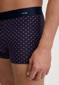 Boxer shorts in dark purple fabric with a geometric diamond pattern. Features a solid navy elastic waistband with "CALIDA" logo.