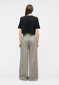 Mujer con cabello largo y castaño, vestida con un top corto negro de manga corta y pantalones grises de pierna ancha y cintura alta, de pie de espaldas a la cámara.