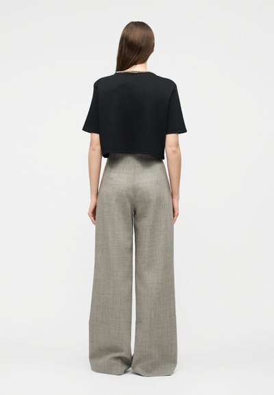 Γυναίκα με μακριά καστανά μαλλιά που φοράει ένα μαύρο κοντομάνικο cropped τοπ και γκρι παντελόνι ψηλόμεσο με φαρδιά μπατζάκια, στέκεται με την πλάτη στην κάμερα.