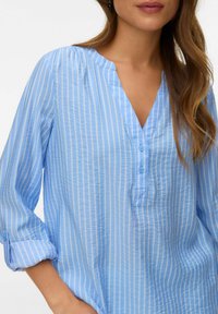 Femme portant un chemisier bleu clair à fines rayures blanches verticales, manches retroussées, et un col en V avec deux boutons.