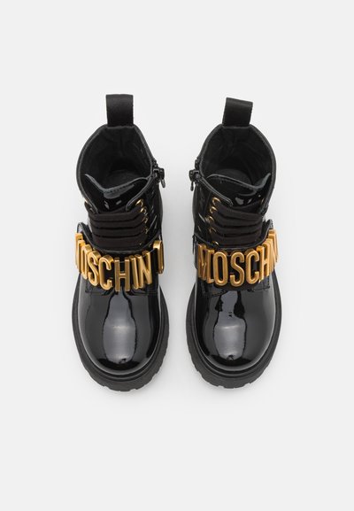 Bottines en cuir verni noir avec une semelle épaisse, présentant des lettres dorées "MOSCHINO" sur la sangle et les lacets, et une fermeture éclair sur le côté.