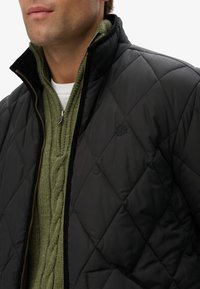 Chaqueta de plumas negra acolchada con un alto cuello, cierre de cremallera, y un suéter verde de punto debajo. Detalle de logo sutil visible.