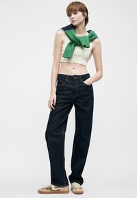 Ženska, oblečena v temnozeleno jeans hlače, bež crop top, superge s leopardjim vzorcem in zeleno jopico, ki jo nosi čez ramena, stoji pred belo ozadjem.