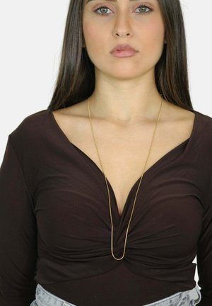 Jeune femme avec des cheveux raides et foncés, portant un haut V à encolure froncée marron foncé et un long collier en or fin.