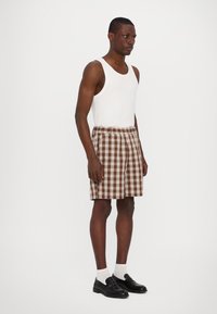 Brown plaid shorts with white and beige accents, elastic waistband. Worn with a white tank top, gray socks, and black loafers.