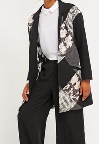 Blazer noir en patchwork avec des motifs floraux et géométriques, en matériaux mixtes, au design sans col et à l'avant ouvert. Porté sur une chemise blanche.
