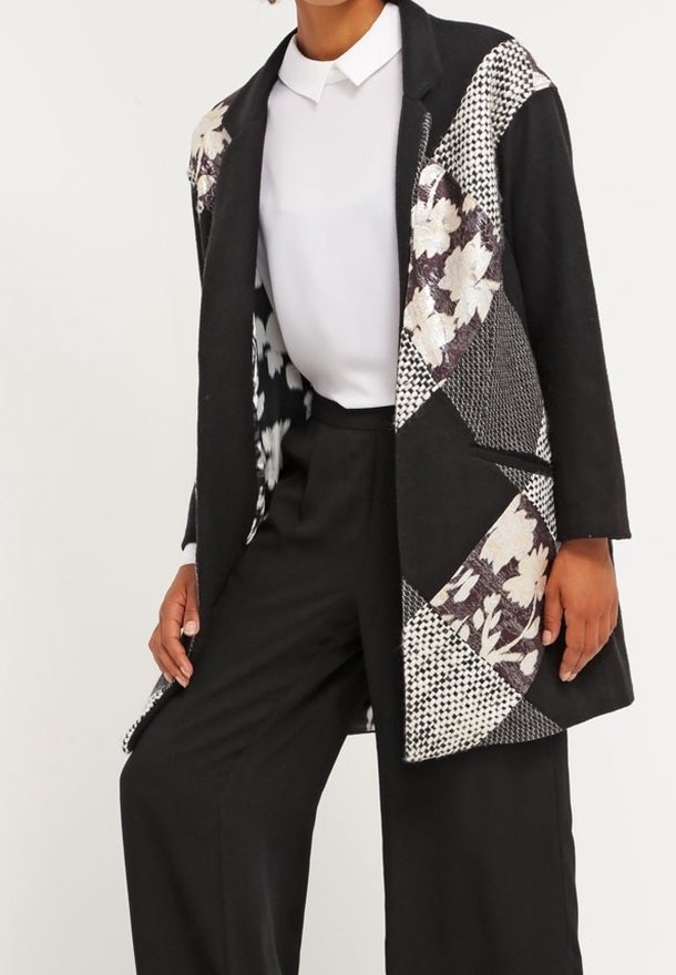 Blazer noir en patchwork avec des motifs floraux et géométriques, en matériaux mixtes, au design sans col et à l'avant ouvert. Porté sur une chemise blanche.