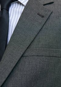 Tamno siva odijak s teksturiranim završetkom, s reverima u obliku slova V i džepom na prsima. Ispod se nalazi košulja s laganim prugama i crna kravata.