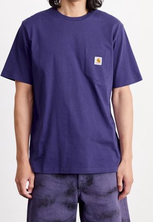 Persona que lleva una camiseta morada lisa de manga corta con un bolsillo en el pecho que presenta un pequeño parche con el logo de Carhartt.