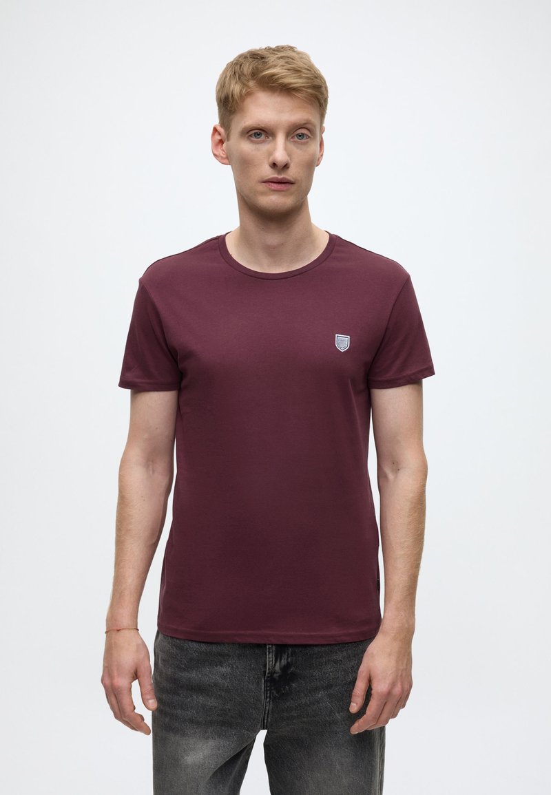 Burgundiapunane puuvillane ümara kaelusega T-särk, mille rinnal on väike kilbi kujuline logo, kombineeritud tumehallide teksadega.