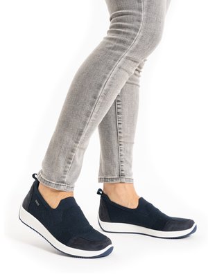 Legs wearing light gray fitted jeans and navy blue slip-on sneakers with white soles standing against a white background.