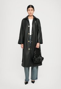 Impermeabile nero in vinile con grandi bottoni dorati, vestibilità oversize e maniche ampie. Abbinato a jeans in denim, décolleté a punta e una piccola borsa nera.