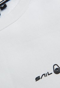 Camiseta blanca con cuello redondo, hecha de una tela suave. Presenta un logo negro de "SAIL" en la esquina inferior derecha.