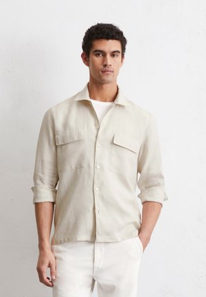 Lys beige linned skjorte med krave, to brystlommer og opkrøllede ærmer. Texturen ser blød og let krøllet ud.
