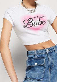 Vit kort t-shirt med texten "not your Babe" i fet svart skrift på en rosa gradientbakgrund. Stylen kombineras med blå högmidjade jeansshorts.