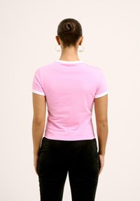 T-shirt ajusté rose avec des bordures blanches au col et aux manches, fabriqué en tissu lisse. Porté avec un pantalon noir, présenté de dos.
