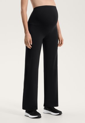 Pantaloni maternità neri con un'ampia fascia elastica in vita, gambe larghe e una texture del tessuto elegante. Abbinati a scarpe sportive nere.