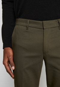 Pantaloni verde oliva realizzati in tessuto morbido, con taglio classico, passanti per cintura e una tasca frontale, abbinati a un maglione nero con maniche.