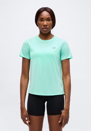 Frau, die nach vorne schaut, trägt ein mintgrünes kurzärmeliges Sportshirt und schwarze enganliegende Shorts vor einem einfarbigen Hintergrund.