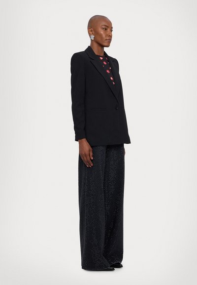 Black tailored blazer with notched lapels over a red patterned top. Loose, sparkly black wide-leg trousers. Silver earrings add detail.