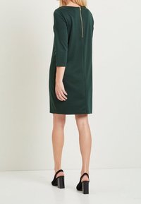 Kvinna klädd i en knälång mörkgrön klänning med trekvartslånga ärmar och dragkedja bak, tillsammans med svarta klackskor med blockklack.