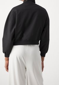 Chaqueta negra recortada con cuello alto y mangas anchas, con una textura suave y puños acanalados; combinada con pantalones claros plisados.