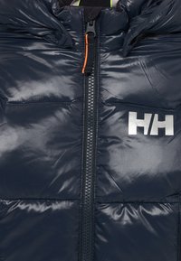 Närbild av en mörkblå vadderad jacka med en central dragkedja och en vit "HH"-logotyp på högra sidan.