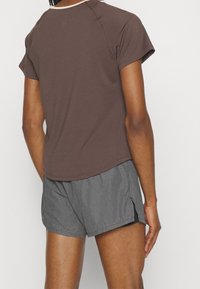 Person som bär en brun kortärmad topp och grå atletiska shorts, står och vänder ryggen mot en enfärgad bakgrund.