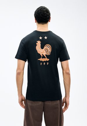 Mann trägt ein schwarzes T-Shirt mit einem stilisierten Hahn, zwei Sternen darüber und "FFF" darunter auf der Rückseite.