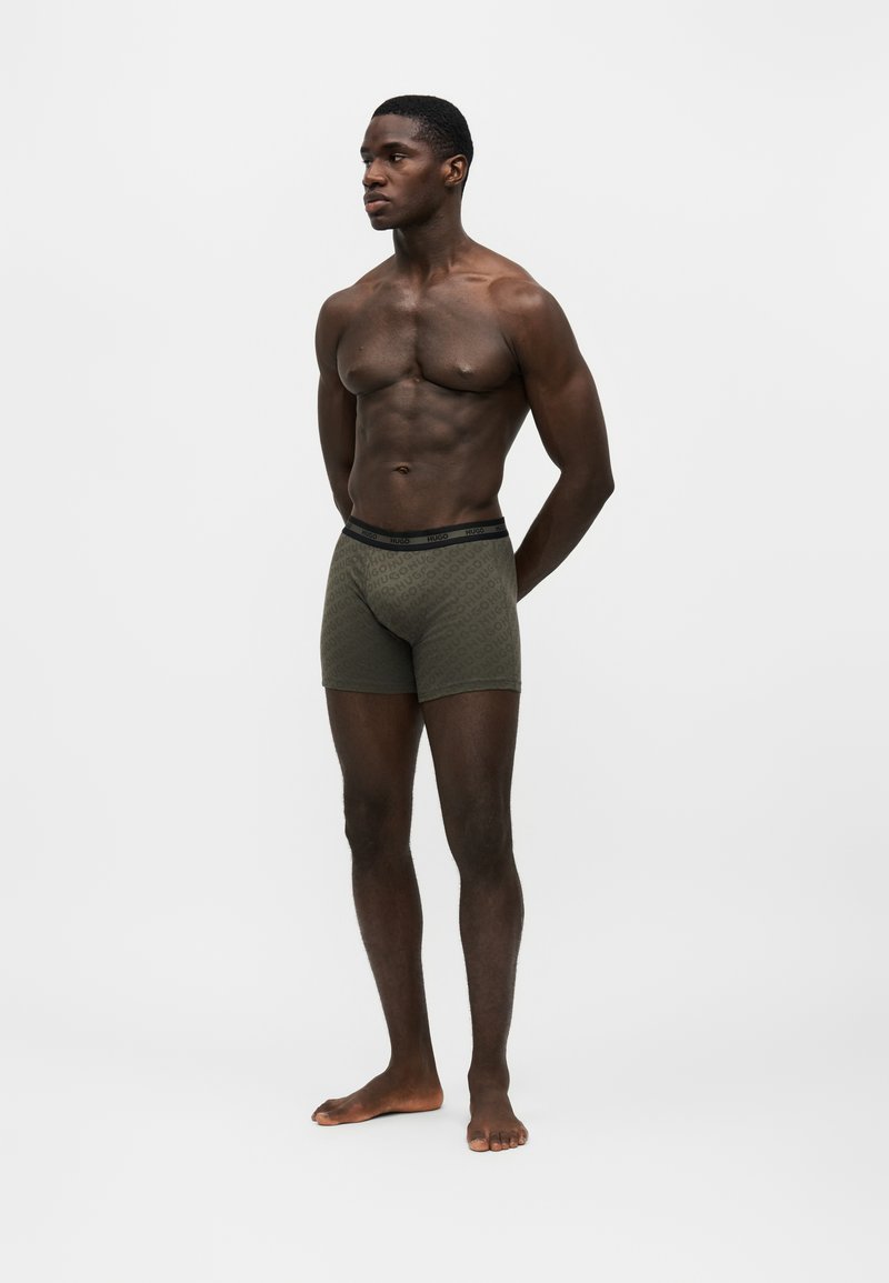 Homme à la peau foncée en forme, debout pieds nus, portant un boxer vert à motifs avec une ceinture noire sur un fond blanc uni.