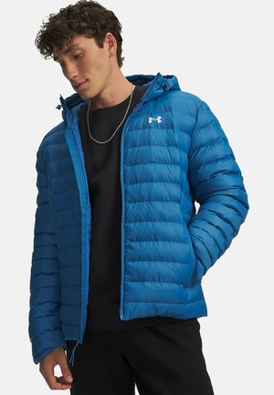 Man die een blauwe donsjacket draagt over een zwart shirt en een zilveren ketting, naar beneden kijkt met één hand in zijn zak.