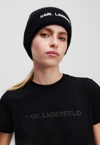 Gorro de punto de ribete negro con el logotipo bordado en blanco "KARL LAGERFELD". Combinado con una camiseta negra que presenta un logotipo texturizado en un estilo similar.