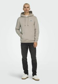 Hoodie beige chiaro con tasca frontale, cordini e patch con logo, abbinato a jeans grigi scuri e scarpe bianche con suola beige.