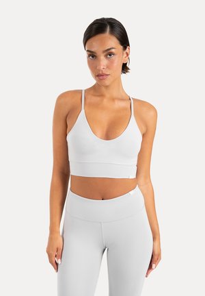 Kvinde iført hvid sports-bh og høj talje leggings, stående mod en ensfarvet lys baggrund, vendt fremad med neutralt udtryk.