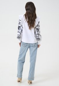 Weißes Blusenshirt mit schwarzen floralen gemusterten Ärmeln und Rüschendetails an den Schultern, kombiniert mit hellblauen Weitbeinjeans und beigen Pumps.