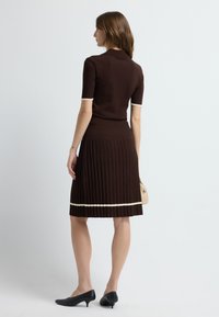 Bruine gebreide jurk met een aansluitend bovenstuk en geplooide rok, met crèmekleurige rand aan de mouwen en zoom. Gemonteerd met zwarte schoenen met hak.