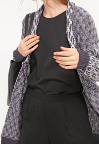 Pletený fialový cardigan s vzorovaným lemem, nošený přes černou blůzu a kalhoty s vysokým pasem, s texturovanou vazbou a otevřeným předním dílem.