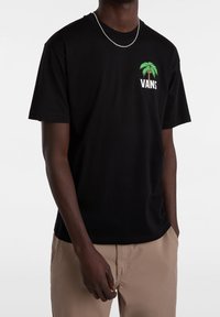 Camiseta de algodón negra con un gráfico de palma verde y el texto "VANS". Mangas cortas, corte relajado, textura suave. Llevada con pantalones beige.