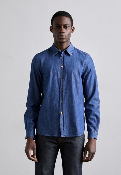 Chemise en denim bleu à manches longues, devant à boutons avec des boutons multicolores, col classique et ourlet droit. Portée avec un jean foncé.