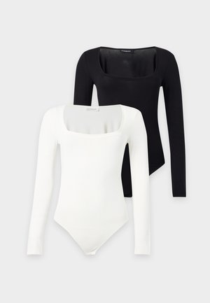 Femme aux cheveux bruns et lisses portant un haut blanc à manches longues, un pantalon noir large à taille haute et des talons noirs, debout devant un fond blanc.