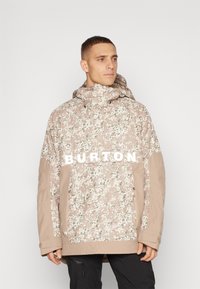 Beige camouflaged jakke med snøre i hætten, lynlåslommer og "BURTON" logo på brystet. Materialet virker struktureret og isoleret.