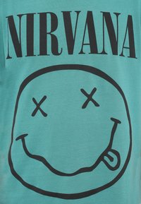 Μπλούζα από βαμβάκι σε τιρκουάζ χρώμα με μεγάλο μαύρο χαμόγελο και το κείμενο «NIRVANA» σε έντονα μαύρα γράμματα από πάνω.