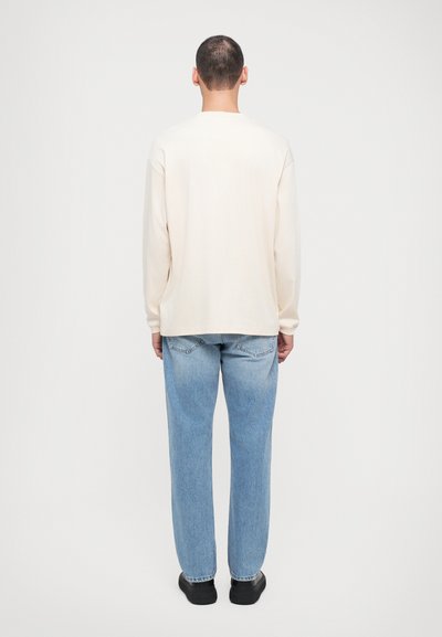 Light beige long-sleeve shirt with dropped shoulders, paired with light blue straight-leg jeans and black shoes, viewed from the back.