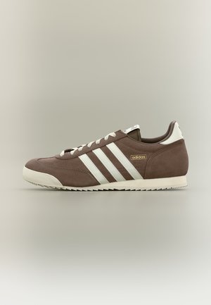 Bruine Adidas lage sneaker met witte strepen, witte zool en veters, zijaanzicht op een neutrale achtergrond.