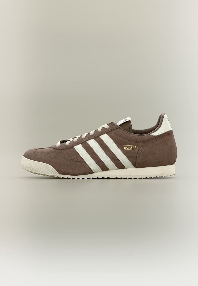 Baskets basses Adidas marron avec bandes blanches, semelle blanche et lacets, montrées de profil sur un fond neutre.